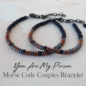 May include: Two black and copper beaded bracelets with a silver clasp. The bracelets are connected by a silver chain. The text "You Are My Person" and "Morse Code Couples Bracelet" is written below the bracelets.