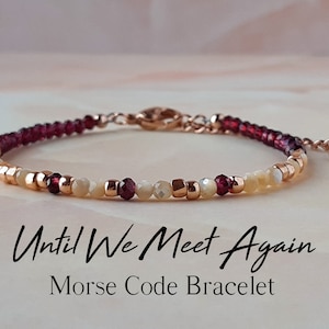 May include: A delicate bracelet with a rose gold chain and beads in shades of red, white, and gold. The bracelet features the message "Until We Meet Again" in Morse code.