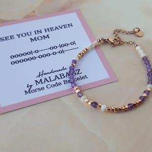 Op de afbeelding: Een paars-witte kralen armband met een ros&eacute;gouden sluiting. De armband heeft de boodschap "See You In Heaven Mom" in morsecode op een roze kaart.