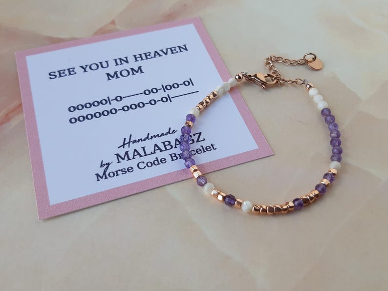 Op de afbeelding: Een paars-witte kralen armband met een ros&eacute;gouden sluiting. De armband heeft de boodschap "See You In Heaven Mom" in morsecode op een roze kaart.