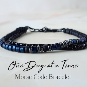 May include: A black and blue beaded bracelet with a silver clasp. The bracelet has the text "One Day at a Time" in cursive writing and the words "Morse Code Bracelet" below it.