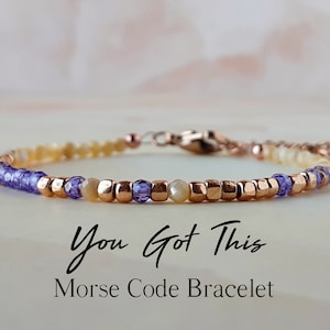 May include: A delicate bracelet featuring a Morse code design. The bracelet combines small, square, rose-gold-colored beads with lavender and pale yellow beads. The text "You Got This" and "Morse Code Bracelet" are displayed in black script.