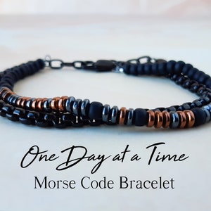May include: A black, gray, and copper beaded bracelet with a metal clasp. The bracelet has the words "One Day at a Time" in cursive script and the words "Morse Code Bracelet" in all caps.