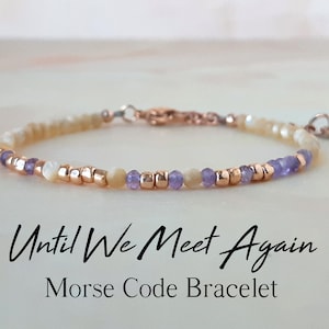 May include: A delicate bracelet with a message in Morse code. The bracelet features small, round beads in shades of white, purple, and gold. The text "Until We Meet Again Morse Code Bracelet" is displayed below the bracelet.