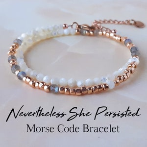 Peut inclure: Un bracelet délicat composé de perles blanches et grises, entrecoupées de petites perles dorées. Le bracelet est doté d'un fermoir en or rose et d'un petit charme. Le bracelet est posé sur une surface blanche et le texte "Nevertheless She Persisted Morse Code Bracelet" est situé en dessous.