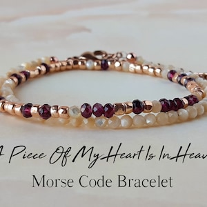 Puede incluir: Pulsera delicada de dos hilos. Uno presenta pequeñas cuentas color crema, mientras que el otro tiene cuentas rojo oscuro y detalles en oro rosa. El texto "A Piece Of My Heart Is In Heaven" y "Morse Code Bracelet" es visible.