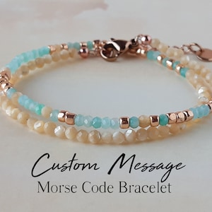May include: A double-strand bracelet with light blue and cream-colored beads and rose gold accents. The bracelet has a clasp and a small charm. The text "Custom Message Morse Code Bracelet" is written below the bracelet.