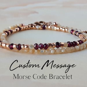 May include: A delicate bracelet with a custom Morse code message. The bracelet features two strands of small, cream-coloured beads, interspersed with rose gold-coloured metal beads and deep red gemstones. The text "Custom Message Morse Code Bracelet" is visible.