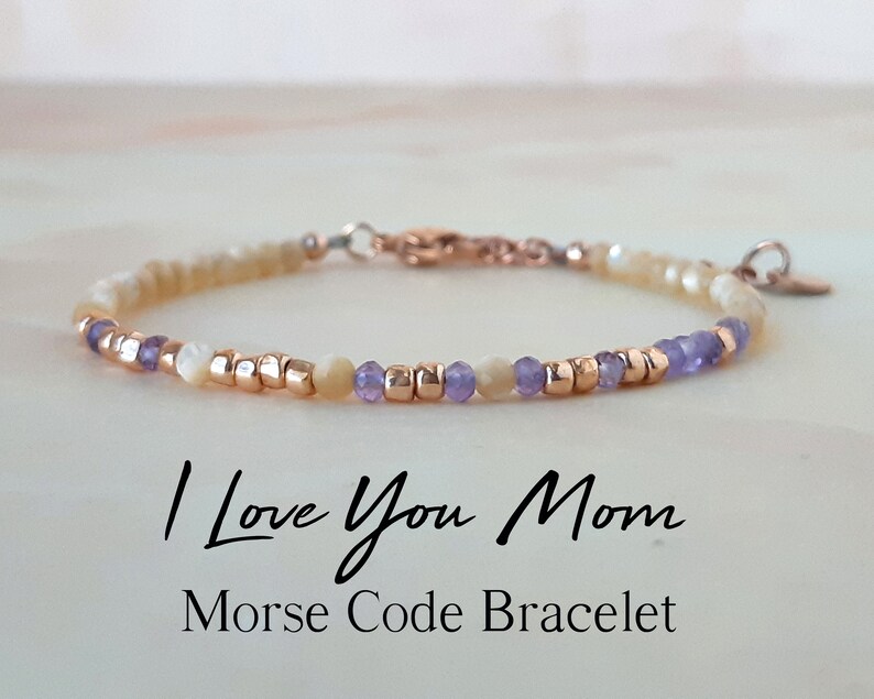 K&ouml;nnte beinhalten: Ein zartes Armband mit kleinen wei&szlig;en und violetten Perlen, die mit goldenen Perlen durchsetzt sind. Das Armband hat einen Verschluss und einen kleinen Herzanh&auml;nger. Der Text "I Love You Mom Morse Code Bracelet" steht unter dem Armband.