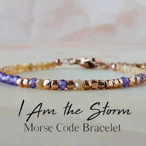 May include: A delicate bracelet with small beads in shades of purple, gold, and white. The bracelet has a clasp closure and features the message "I Am the Storm" in Morse code.