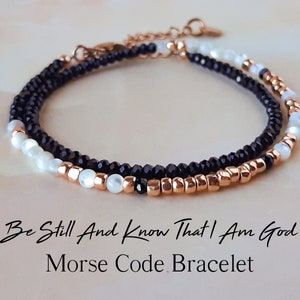 May include: A delicate bracelet with black and white beads and rose gold accents. The bracelet is layered and features a message in Morse code: "Be Still And Know That I Am God".