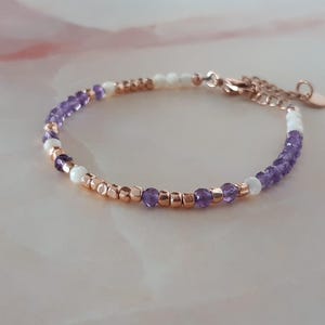 Op de afbeelding: Een delicate armband met een mix van paarse en witte kralen, geaccentueerd met roosgoud kralen en een roosgoud sluiting.