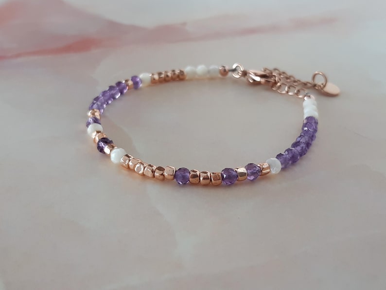 Op de afbeelding: Een delicate armband met een mix van paarse en witte kralen, geaccentueerd met roosgoud kralen en een roosgoud sluiting.