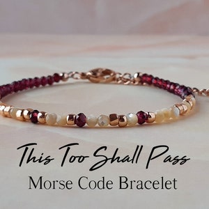 Könnte beinhalten: Ein zartes Armband mit roten und weißen Perlen, mit einem roségoldenen Verschluss und einem Herz-Anhänger. Das Armband trägt die Botschaft "This Too Shall Pass" in Morse-Code.