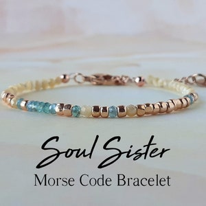 May include: A delicate bracelet featuring white, blue and gold beads. The bracelet has a clasp and a small charm. The text "Soul Sister Morse Code Bracelet" is written below the bracelet.