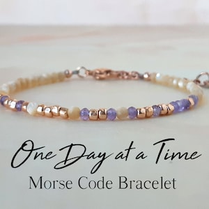 Op de afbeelding: Een delicate armband met een boodschap in morsecode. De armband heeft kleine, ronde kralen in tinten wit, paars en goud. De tekst "One Day at a Time" staat onder de armband.