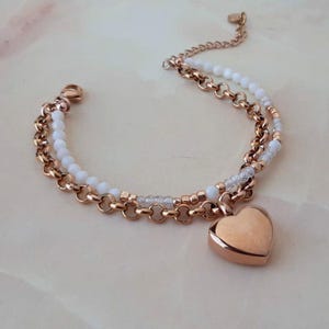 Op de afbeelding: Een dubbelstrengs armband met roségoudkleurige metalen kettingschakels, kleine witte kralen en doorzichtige gefacetteerde kralen. Een gladde, massieve hartvormige bedel in roségoud hangt aan het midden van de armband, voorzien van een karabijnsluiting.