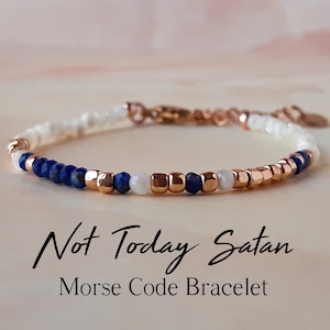 Puede incluir: Un brazalete de cuentas con cuentas blancas, azules y doradas. El brazalete deletrea "Not Today Satan" en código Morse.