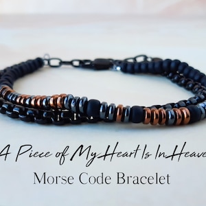 May include: A black, gray, and copper beaded bracelet with a metal clasp. The bracelet has the text "A Piece of My Heart Is In Heaven" and "Morse Code Bracelet" written below it.