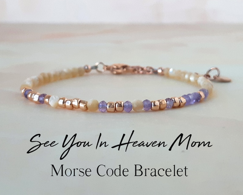 Op de afbeelding: Een delicate armband met kleine witte en paarse kralen en een gouden sluiting. De armband heeft de boodschap "See You In Heaven Mom" in morsecode.