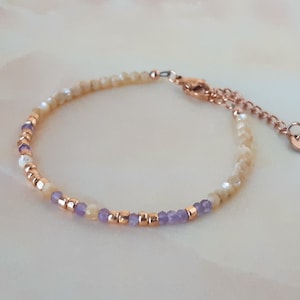 K&ouml;nnte beinhalten: Ein zartes Armband mit einem Mix aus kleinen, schimmernden wei&szlig;en Perlen und lila Perlen. Das Armband hat einen ros&eacute;goldenen Verschluss und eine Kette.