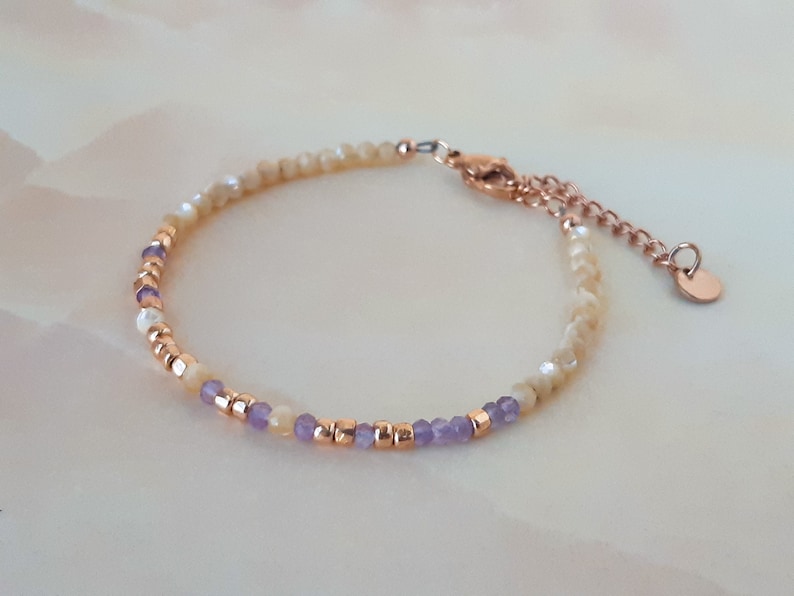 K&ouml;nnte beinhalten: Ein zartes Armband mit einem Mix aus kleinen, schimmernden wei&szlig;en Perlen und lila Perlen. Das Armband hat einen ros&eacute;goldenen Verschluss und eine Kette.