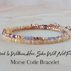 Può includere: Un braccialetto a doppia fila con un design delicato. Il braccialetto presenta piccole perline di colore chiaro e perline viola. Il braccialetto è realizzato con dettagli in oro rosa e ha una chiusura a moschettone. Il braccialetto è accompagnato dal testo "God is Within Her, She Will Not Fail" e "Morse Code Bracelet".