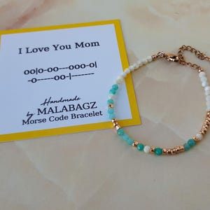 May include: A delicate bracelet with white and turquoise beads and a gold clasp. The bracelet is accompanied by a yellow card with the text "I Love You Mom" and Morse code for the message. Handmade by MALABAGZ. Morse Code Bracelet.