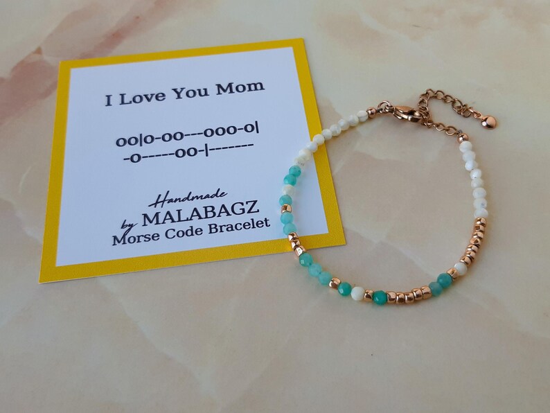 May include: A delicate bracelet with white and turquoise beads and a gold clasp. The bracelet is accompanied by a yellow card with the text "I Love You Mom" and Morse code for the message. Handmade by MALABAGZ. Morse Code Bracelet.