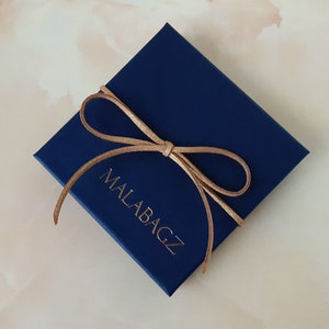 Op de afbeelding: Een donkerblauwe cadeaudoos met een bruin lint dat in een strik is gebonden. Op de doos staat de tekst "MALABAGZ" gedrukt.
