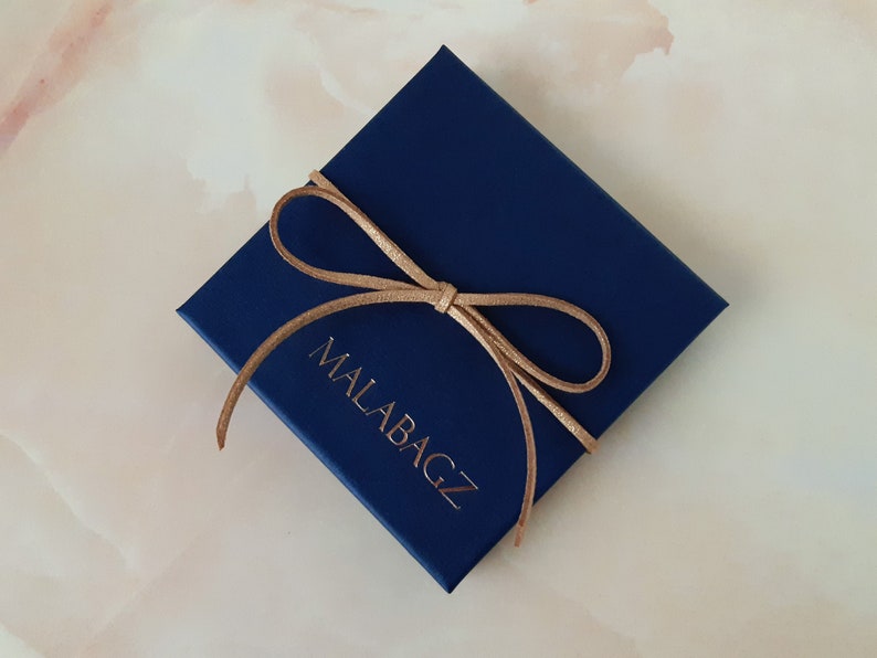 Op de afbeelding: Een donkerblauwe cadeaudoos met een bruin lint dat in een strik is gebonden. Op de doos staat de tekst "MALABAGZ" gedrukt.