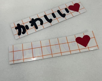 May include: Two rectangular stickers with the Japanese word "kawaii" in black and a red heart. The stickers are on a white background with a grid pattern.