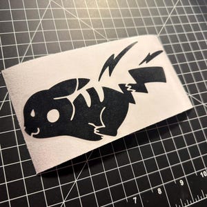 May include: Black vinyl decal of a stylized Pikachu character with lightning bolt details. The decal is on a rectangular, off-white background, set against a black grid surface. The design features a simplified, cartoonish aesthetic.