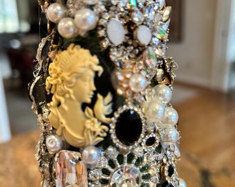 May include: A black bottle covered in a variety of rhinestones, pearls, and other decorative elements. The bottle is decorated with a vintage cameo design.
