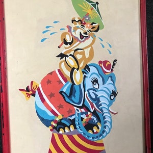 May include: A vintage framed painting of a blue elephant with a red hat and a yellow animal with a green umbrella riding on its back. The elephant is standing on a red and white striped platform with a red and white star-shaped ball.