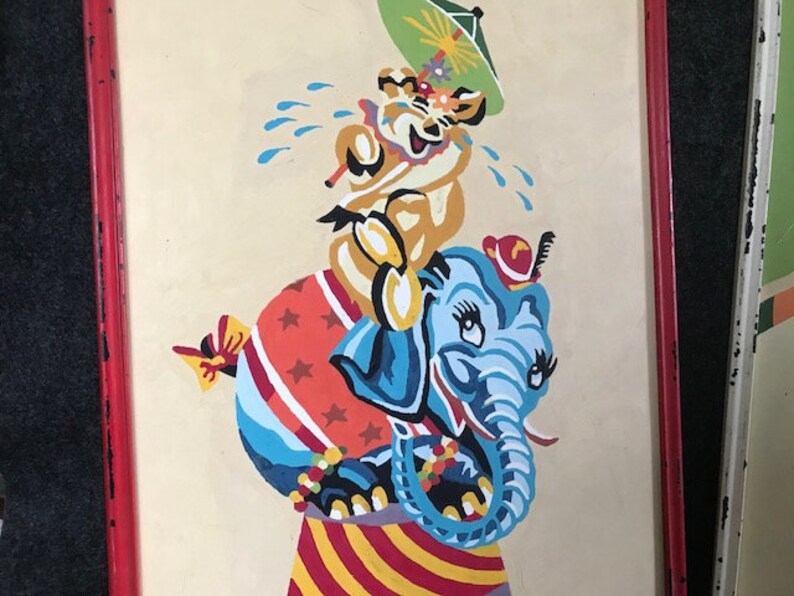 May include: A vintage framed painting of a blue elephant with a red hat and a yellow animal with a green umbrella riding on its back. The elephant is standing on a red and white striped platform with a red and white star-shaped ball.