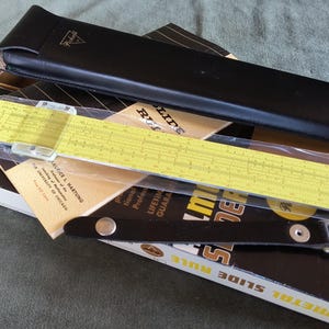 May include: A vintage Pickett slide rule, still in its original packaging, with a yellow and black design. The slide rule is accompanied by a black leather case and a box with the text "Slide Rule".