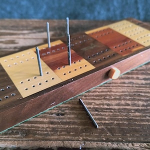 May include: A wooden cribbage board with a light brown, dark brown, and red wood grain pattern. The board has a green felt lining and a small orange button on the side. There are four metal pegs inserted into the board.