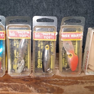 May include: Five fishing lures in their original packaging. The lures are in various colors, including red, blue, silver, and orange. The packaging includes the brand name "Tumble Bug" and the lure names "Wee Wart" and "Hot'n Tot".