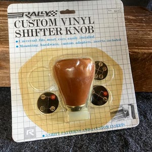 May include: A brown vinyl custom shifter knob with a gold metal base and five different shift pattern inserts. The packaging includes the text "RALLY CUSTOM VINYL SHIFTER KNOB" and "Universal fits most cars easily installed. Mounting hardware, custom adapters, inserts included." The package also includes the number "4625" and the text "5 SHIFT PATTERNS AND CUSTOM INSERTS".