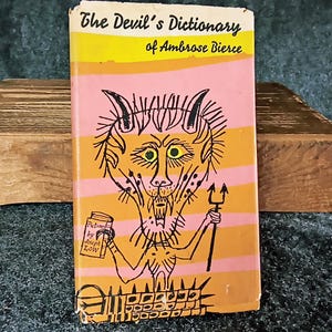 May include: A vintage paperback book titled "The Devil's Dictionary of Ambrose Bierce" with a cartoon illustration of a devil holding a pitchfork and a book.