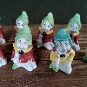 May include: A collection of ceramic figurines depicting small, gnome-like characters. Each figure wears a green hat, a red coat, and a yellow collar. The figurines are arranged on a wooden surface.
