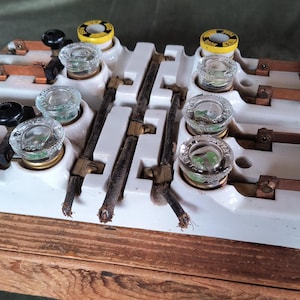 May include: An antique electrical component with white ceramic bases, copper contacts, and glass fuse holders. The device has black knobs and is mounted on a wooden base. The fuses have yellow and black tops. A vintage electrical item.