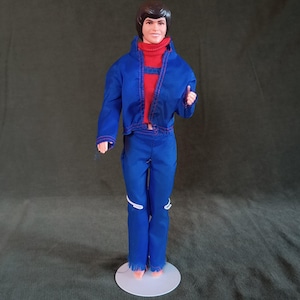 May include: A vintage doll wearing a blue jacket and pants with a red turtleneck sweater. The jacket has red stitching and the pants have white accents. The doll has brown hair and is standing on a white circular base.