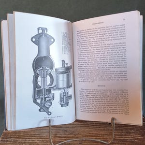 May include: An open book with detailed black and white illustrations of a carburetor. The book is propped open on a metal stand, with text on the right page. The book is on a wooden surface.