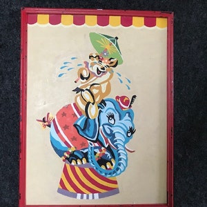 May include: A framed painting of a blue elephant with a red hat and a yellow animal with a green umbrella riding on its back. The elephant is standing on a red and white striped platform.