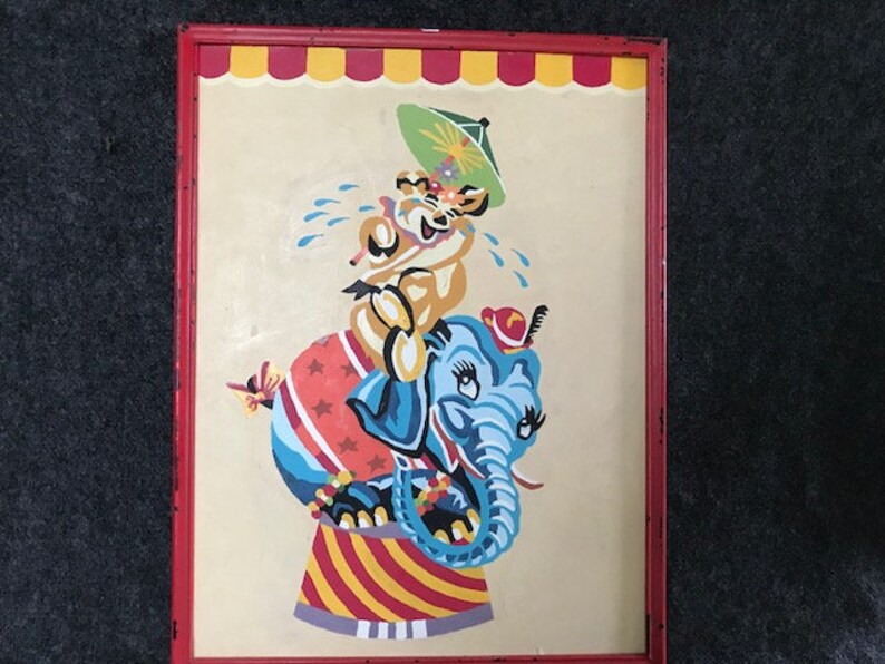 May include: A framed painting of a blue elephant with a red hat and a yellow animal with a green umbrella riding on its back. The elephant is standing on a red and white striped platform.