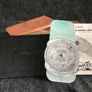 May include: A Weems & Plath Dead Reckoning Computer, a circular slide rule with a green grid, and a black leather flight log book. The computer has a white background with black text and markings. The flight log book has the text "Flight Log" printed on the cover.