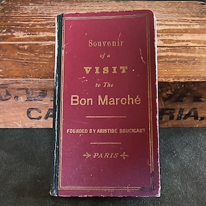 May include: A vintage burgundy-colored booklet titled "Souvenir of a Visit to The Bon Marché" with gold lettering. The booklet is set against a wooden crate background. The text includes "Founded by Aristide Boucicaut" and "Paris".