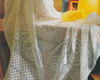 May include: A white, open-weave crochet blanket draped over a table. The blanket features a repeating geometric pattern and is partially folded, revealing the intricate design. A wicker basket and yellow decor are in the background.